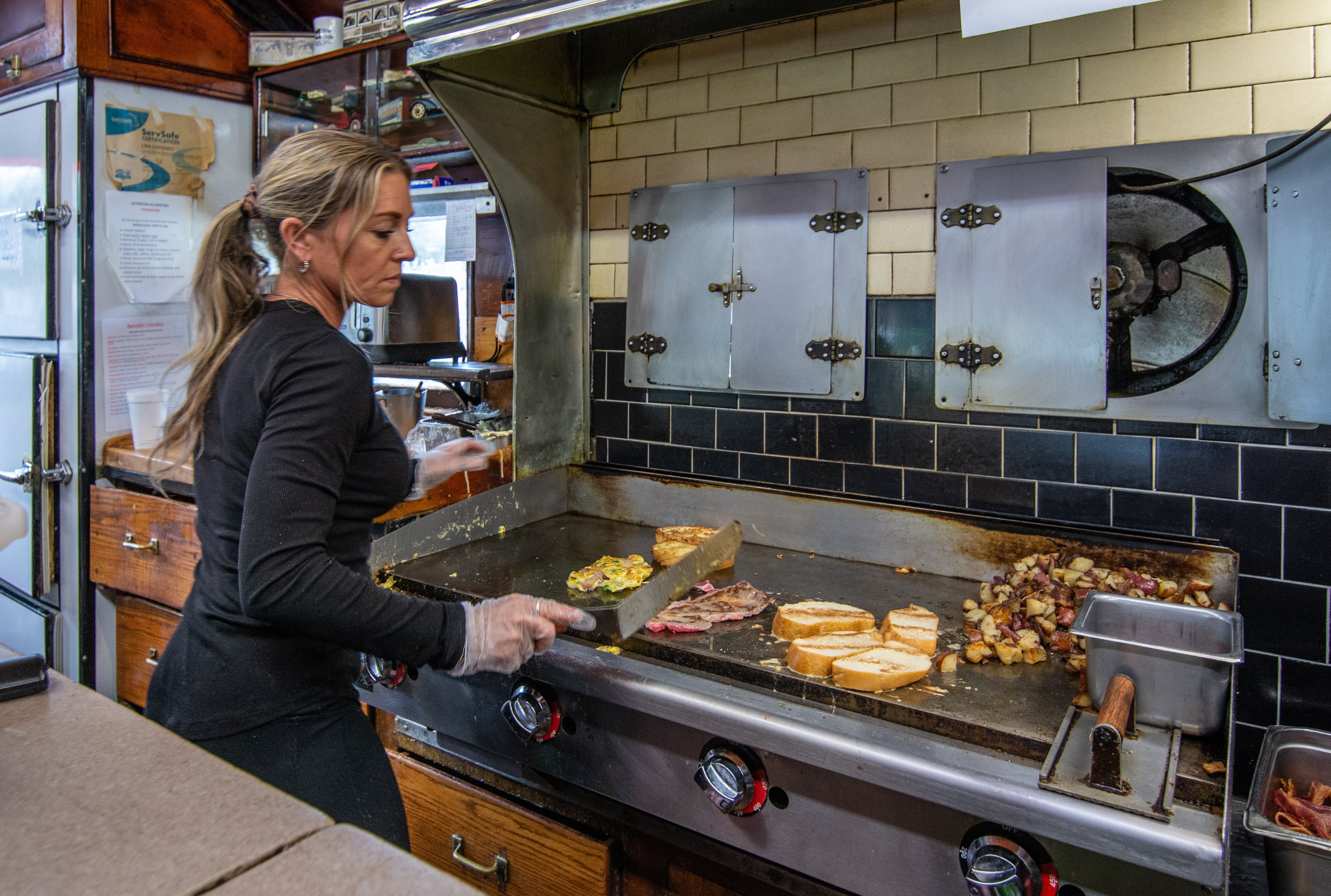 A woman with blonde hair pulled back in a ponytail works at a diner grill with eggs, meat, home fries, and bread frying on it.