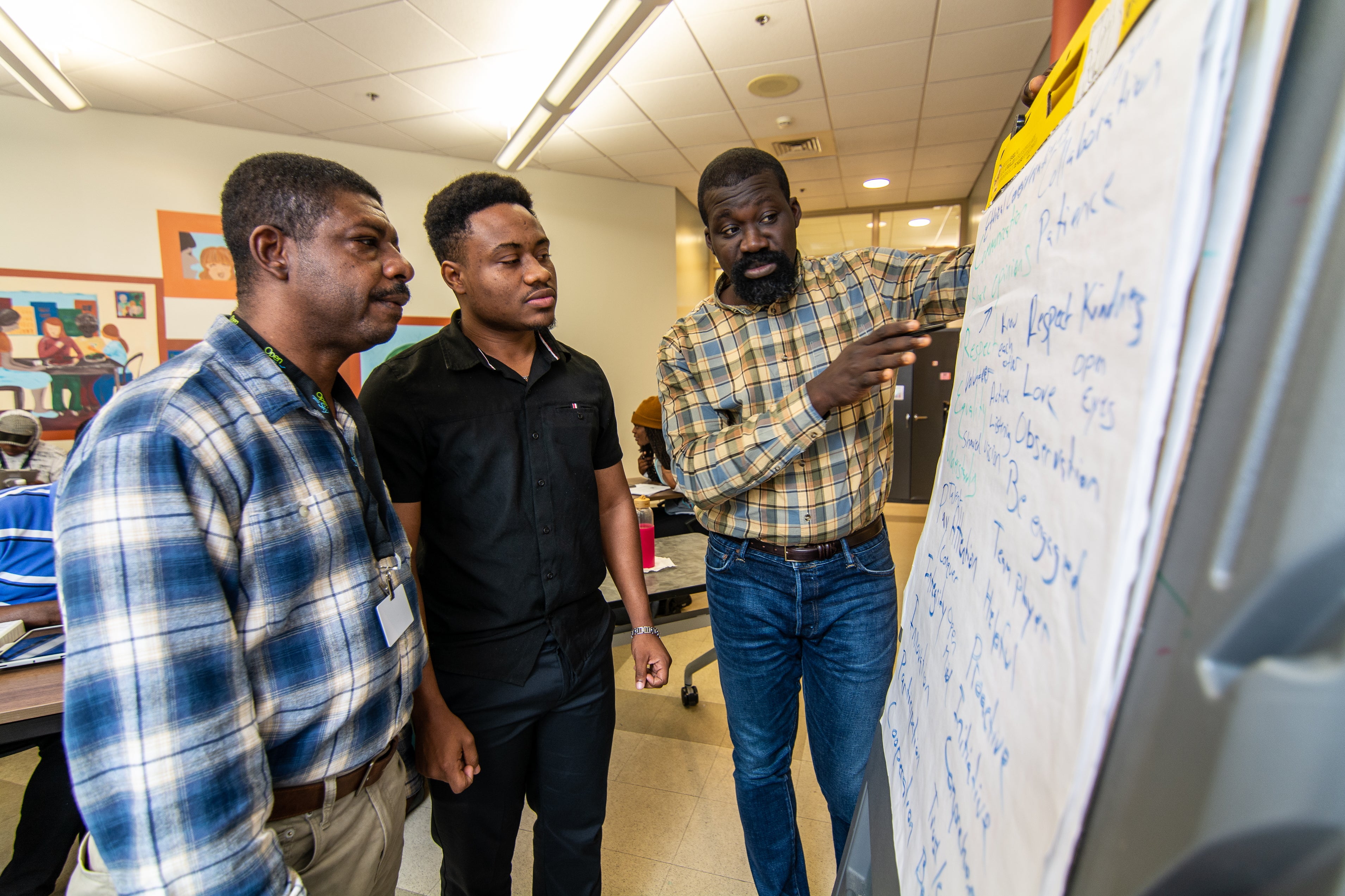 Three men stand in front of an easel pad with writing on it inside a classroom.