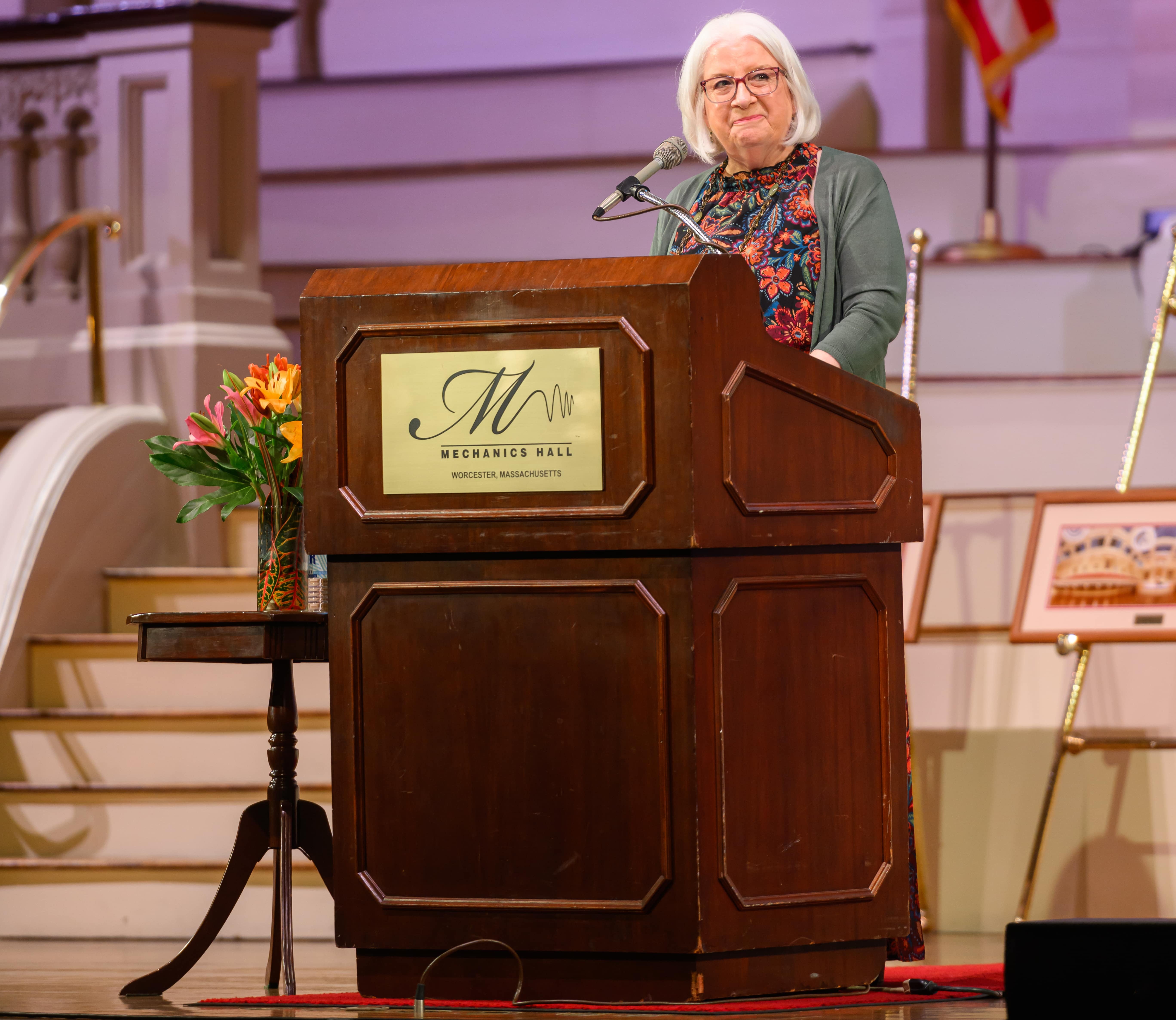 A woman wearing a floral shirt and a gray sweater stands behind a podium at Mechanics Hall.