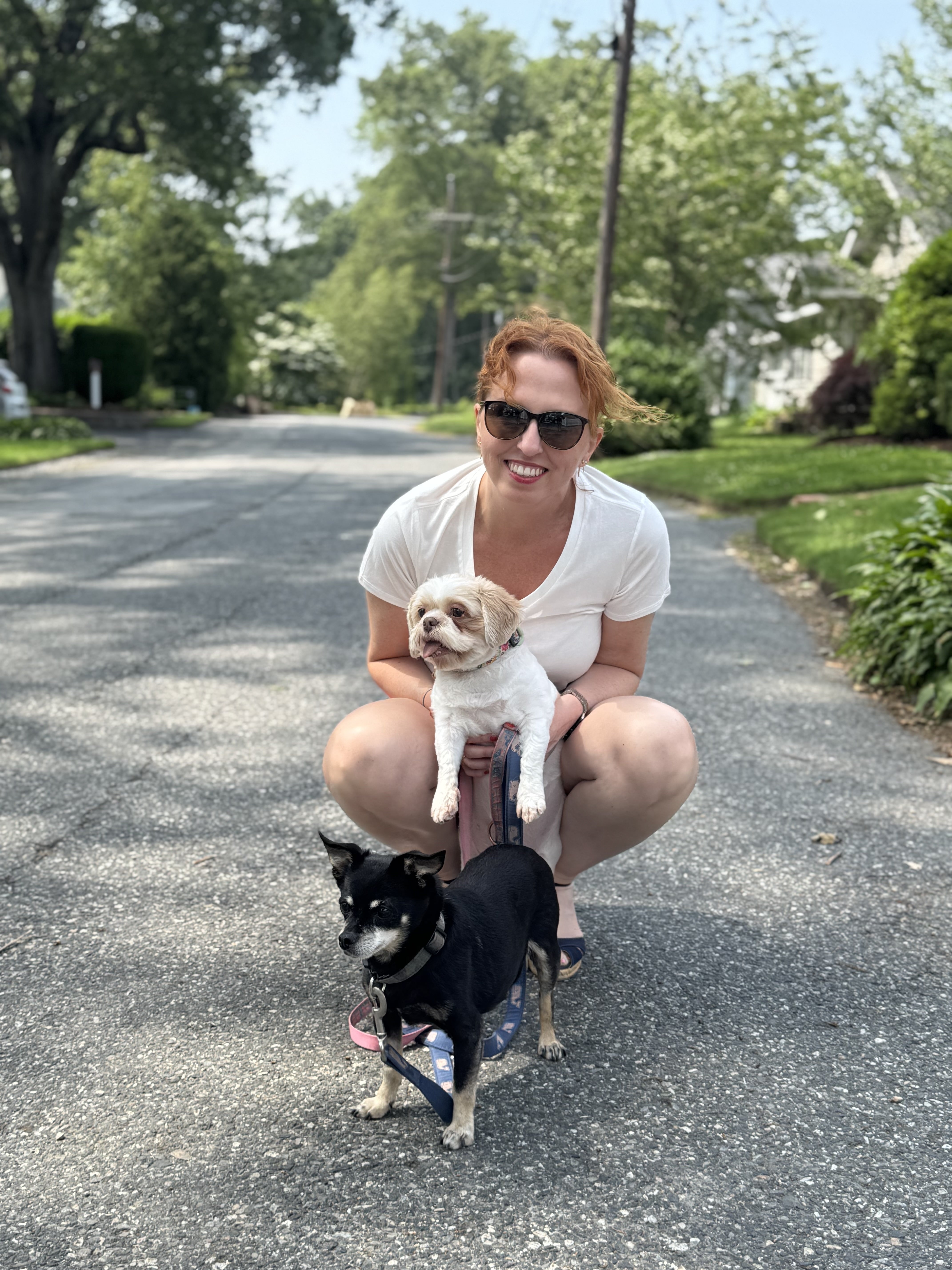 A woman with red hair holds a amll white dog with a small balck dog at her feet.