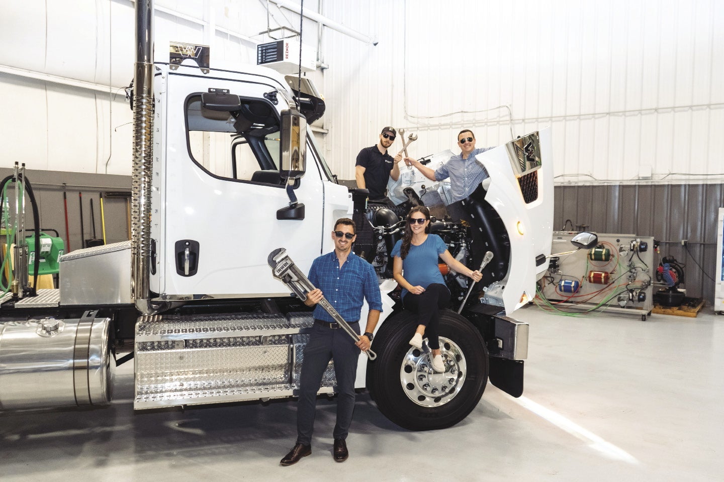Four people holding wrenches while sitting on or around the open hood of a large white truck in a garage