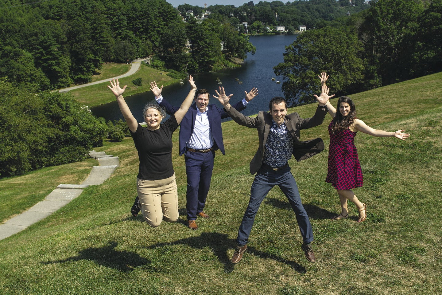 Four people jump in the air on a grassy hill overlooking a lake.