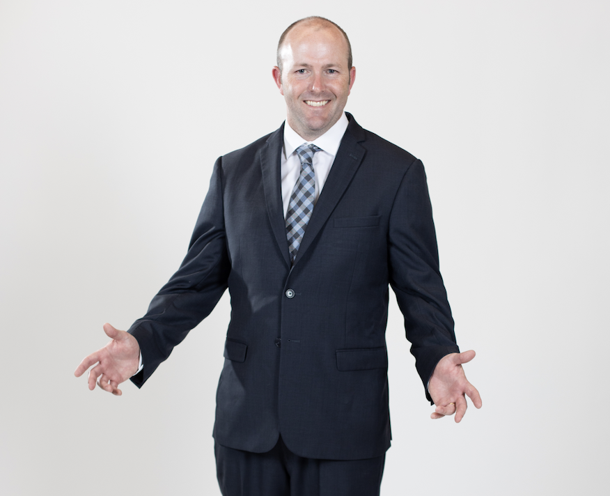 A man wearing a dark blue suit stands with his hands held out beside him.