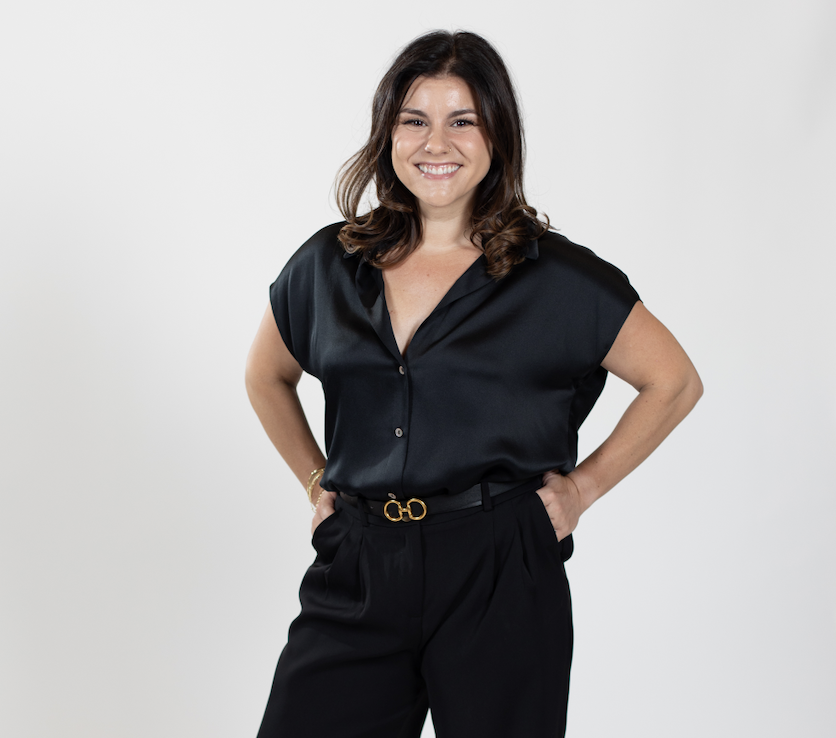 A woman with shoulder length brown hair stands with her hands in her pant pockets wearing a black top and pants.