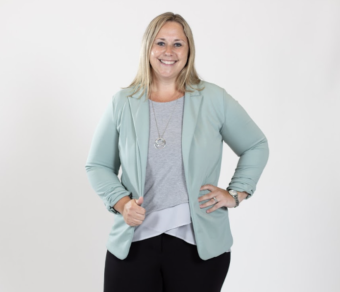 A woman with blonde hair wears a grey bouse and a pale blue blazer.