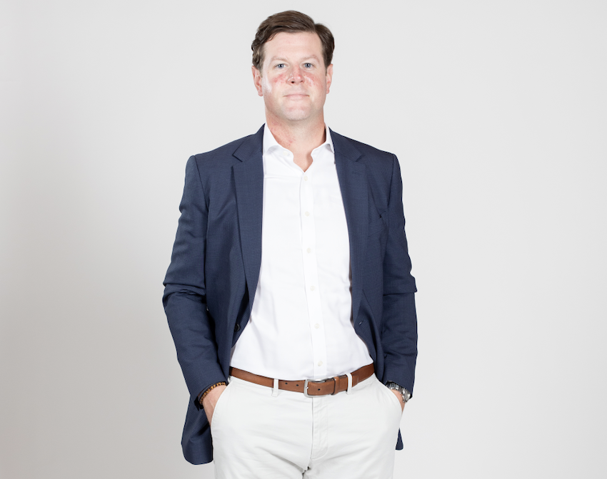 A man stands wearing a white shirt and pants with a blue blazer and his hands tucked into his pockets.