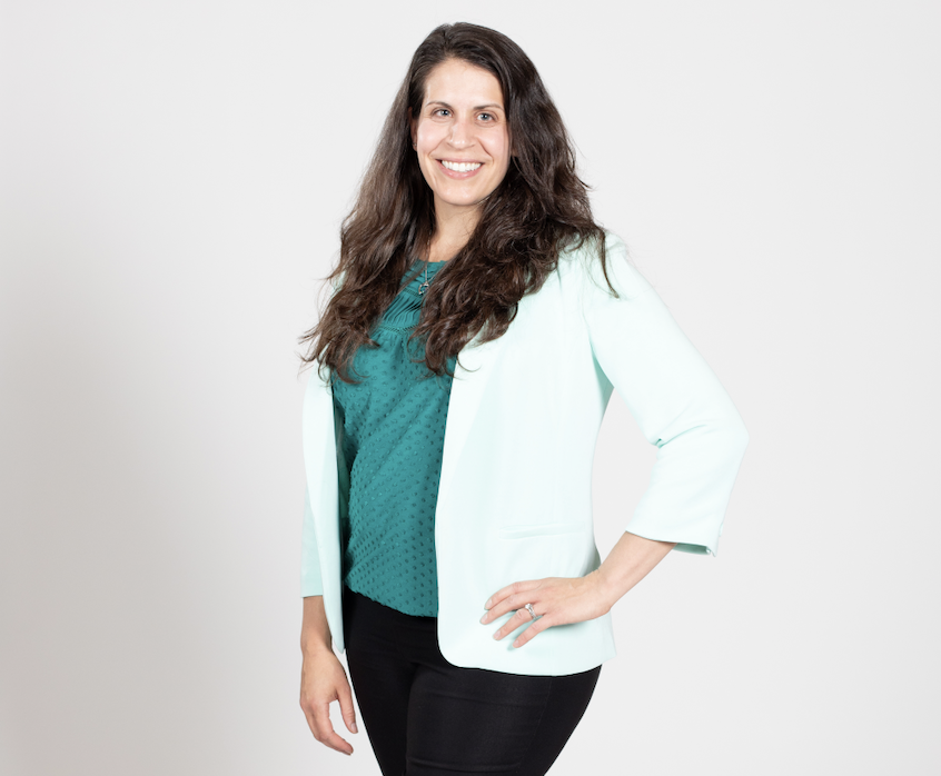 A woman with long brown hair wears a teal blouse, light mint blazer, and black pants