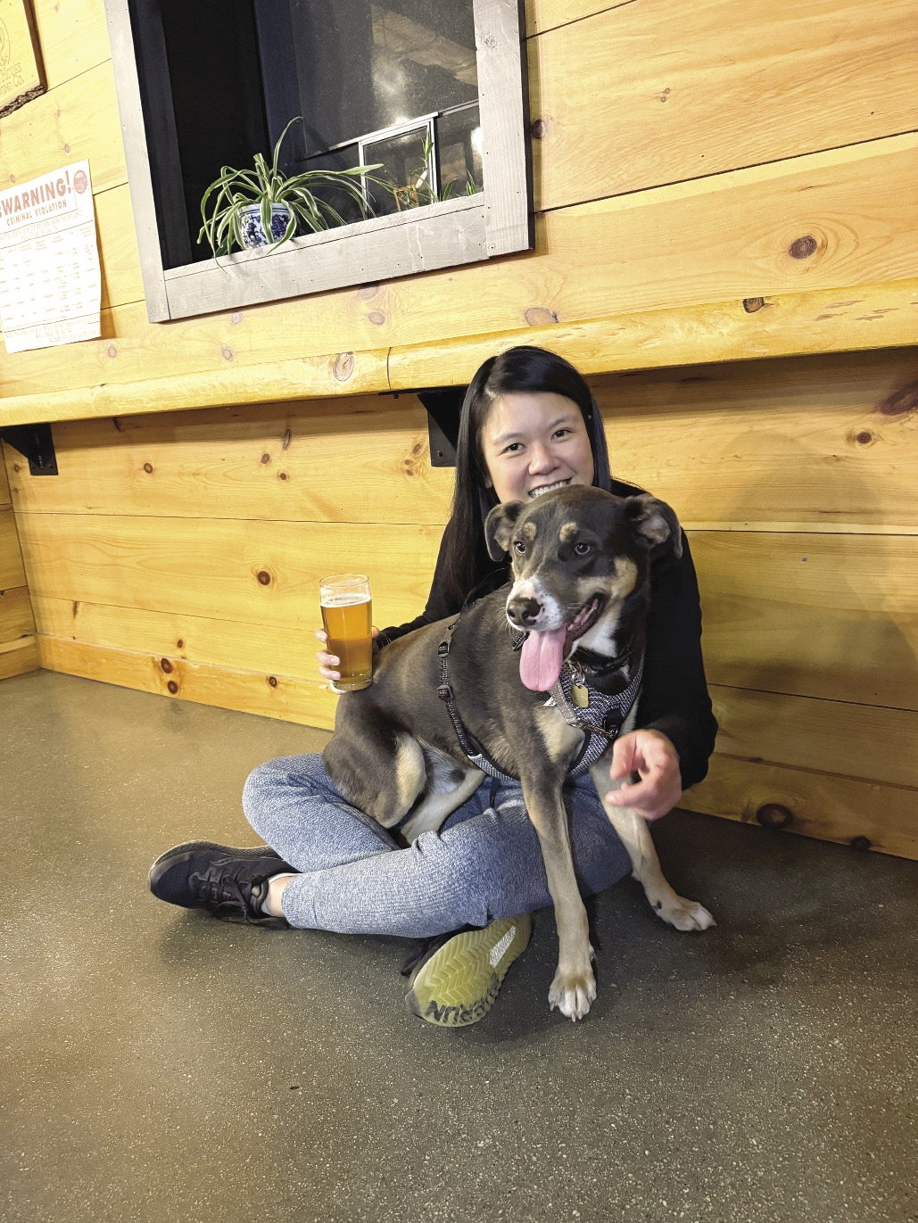 A woman with dark hair holds a beer and sits on the floor as a dog sits on her lap.