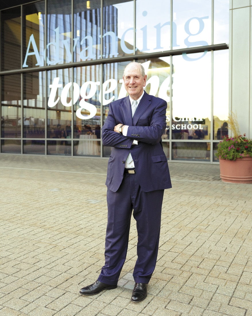 A man stands with his arms crossed while wearing a dark blue suit, standing in front of university building with the words "Advancing together" printed on the windows. 