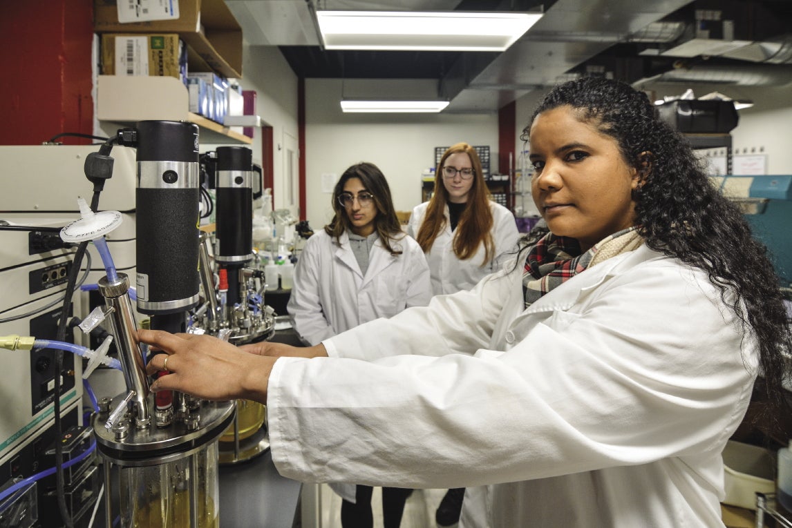 Three women in white lab coats work on research equipment.