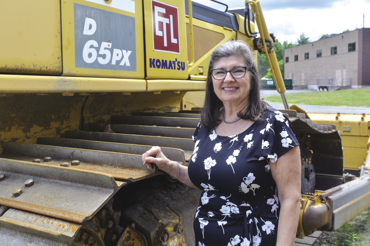 A woman in a black dress with a white, floral pattern stands with her hand on a large yellow front loader.