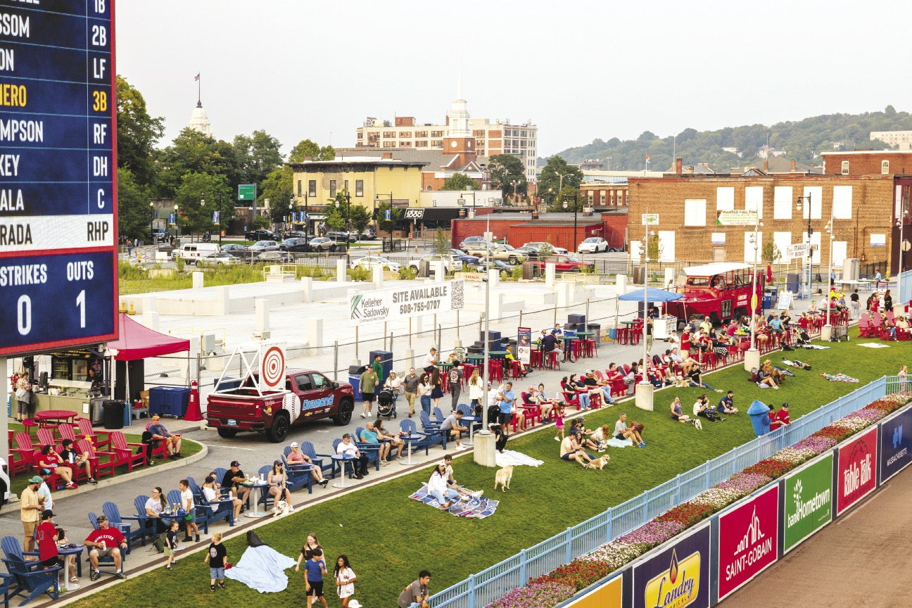 People sit on a grass bern in the left field area of a minor league ballpark with an undeveloped commerical site fenced off behind them. 