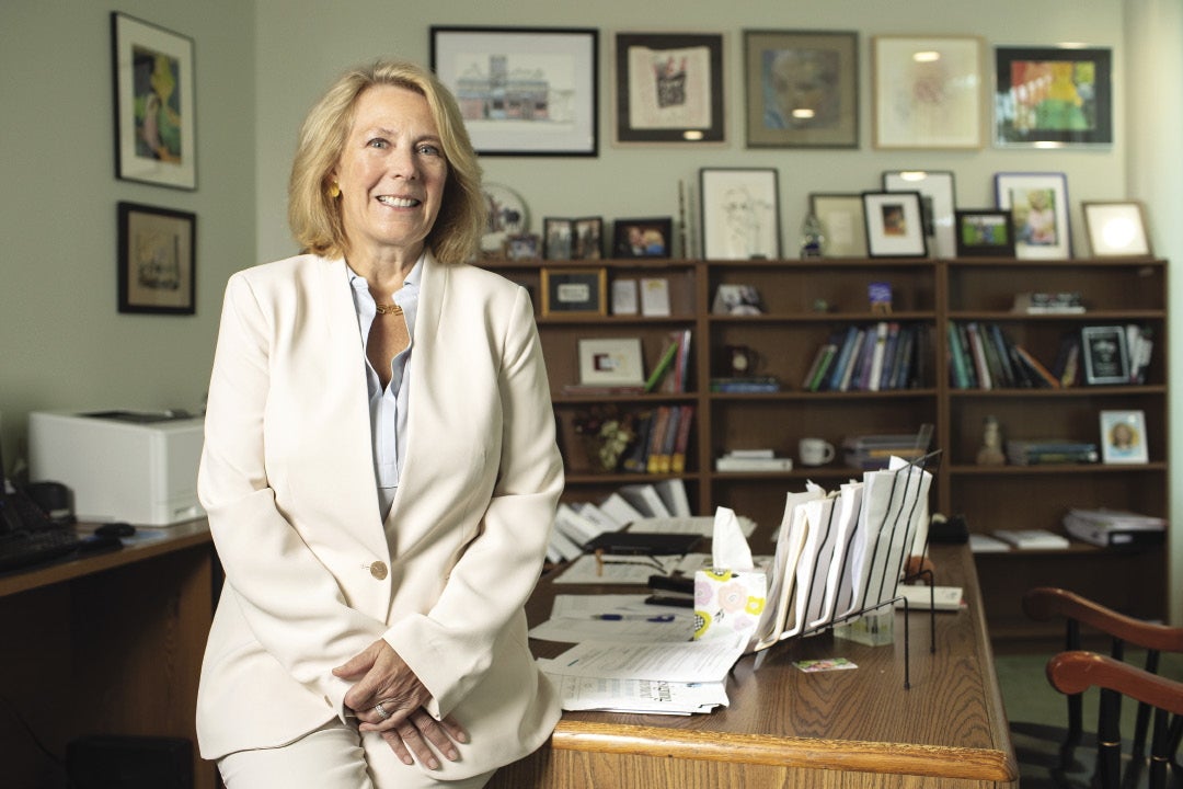 A woman in a white suit leans on her desk in her office.