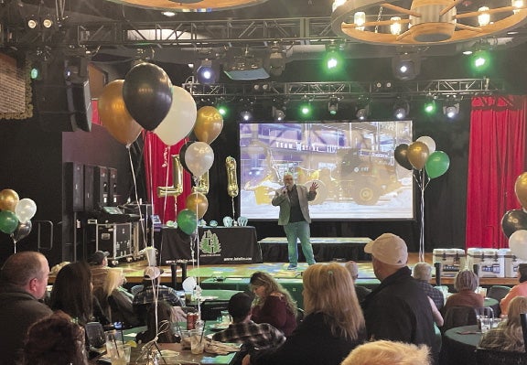 A man stands on a stage in front a crowd seated at tables, with balloons and stage lights visable