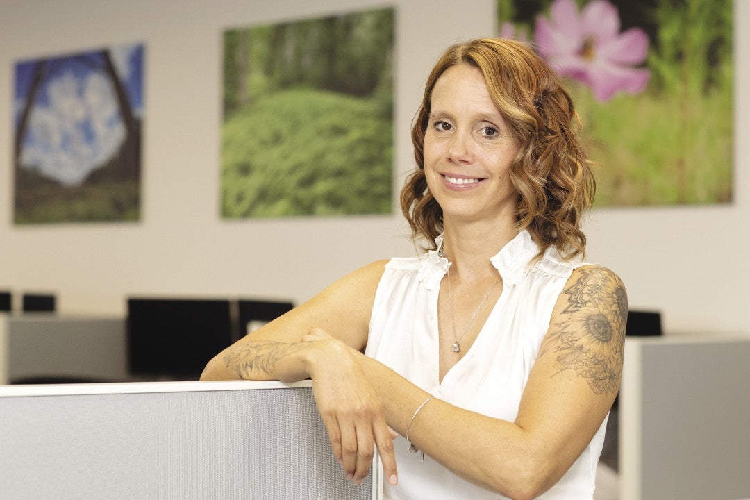 A smiling woman leans on the top of a cubicle wall in an office
