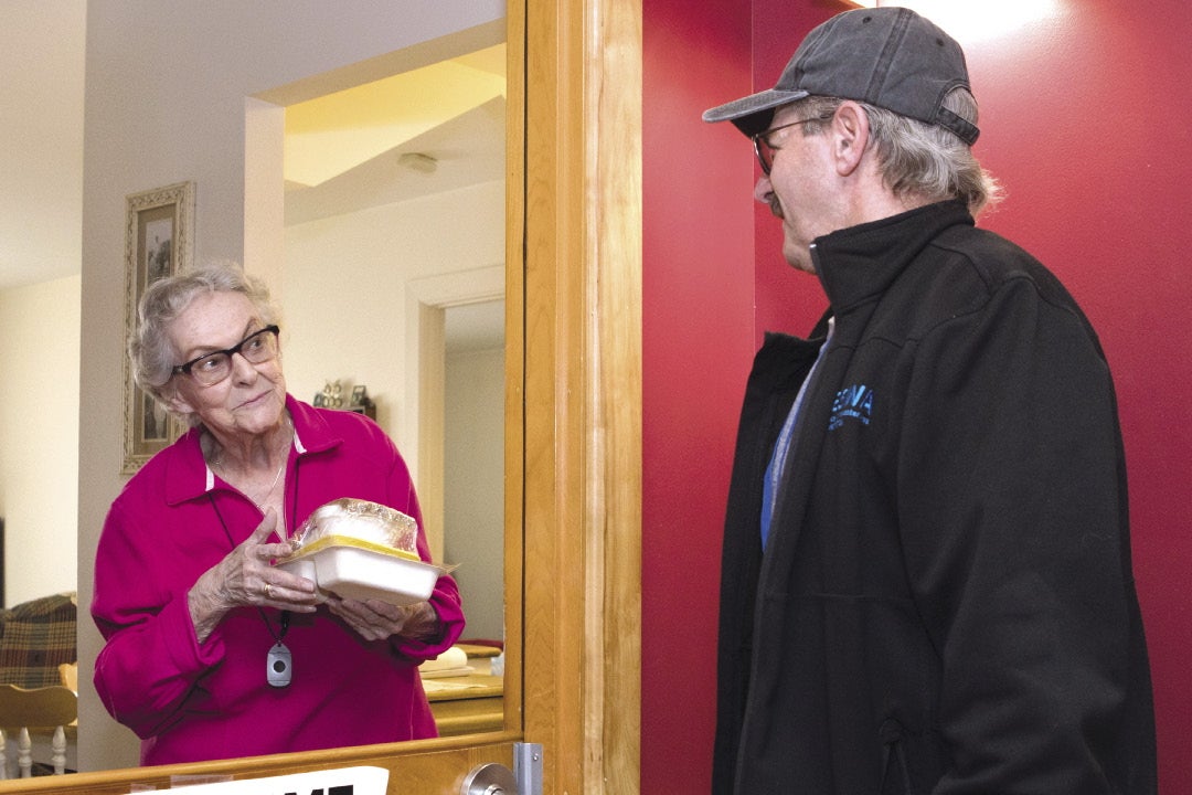 A woman with grey hair and glasses wearing a pink button down holds a couple cartons of food as she looks through a door window at a man with grey hair wearing a grey baseball cap and a black zip up jacket.