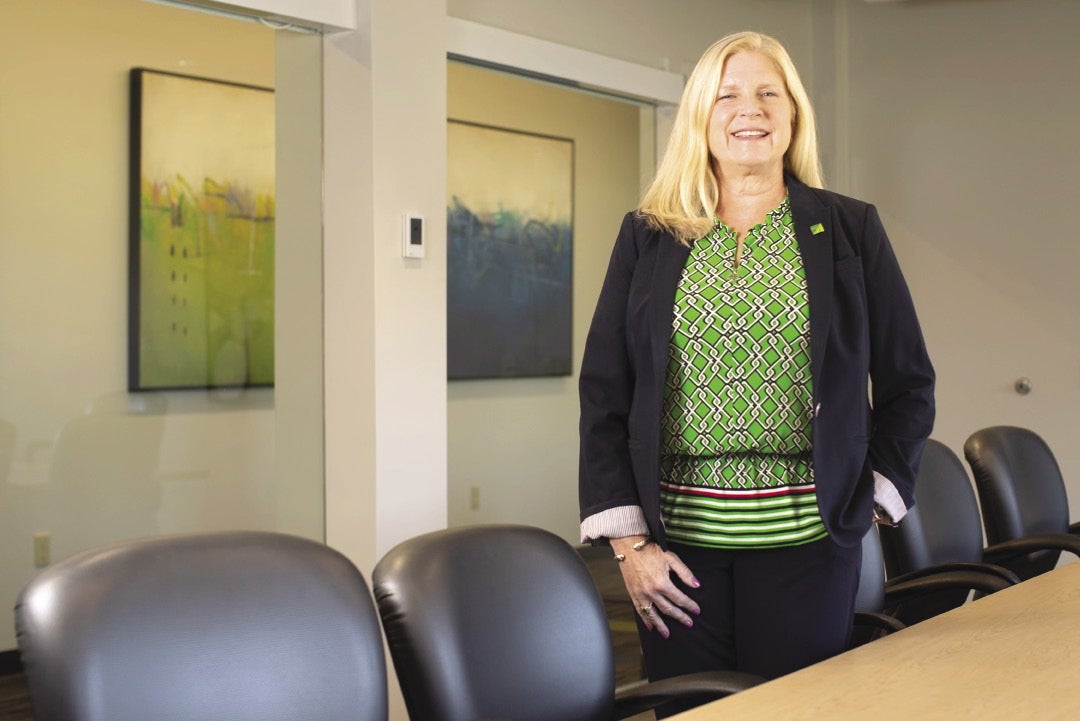 A woman wearing a green shirt and blue blazer stands in a conference room.
