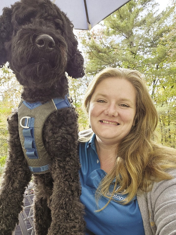 A woman with long blonde hair sits next to a black doodle dog. 
