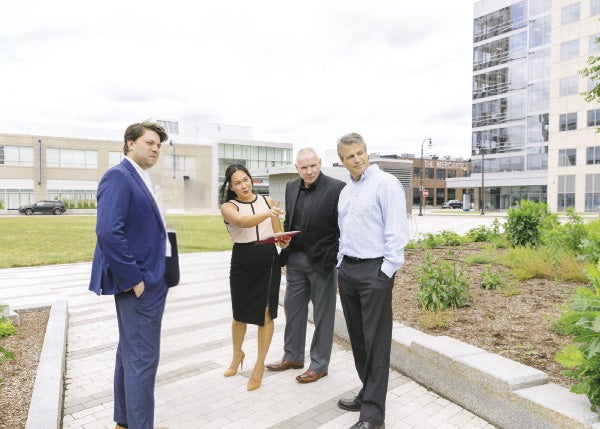Four people stand in the middle of an urban green, looking at something off camera.