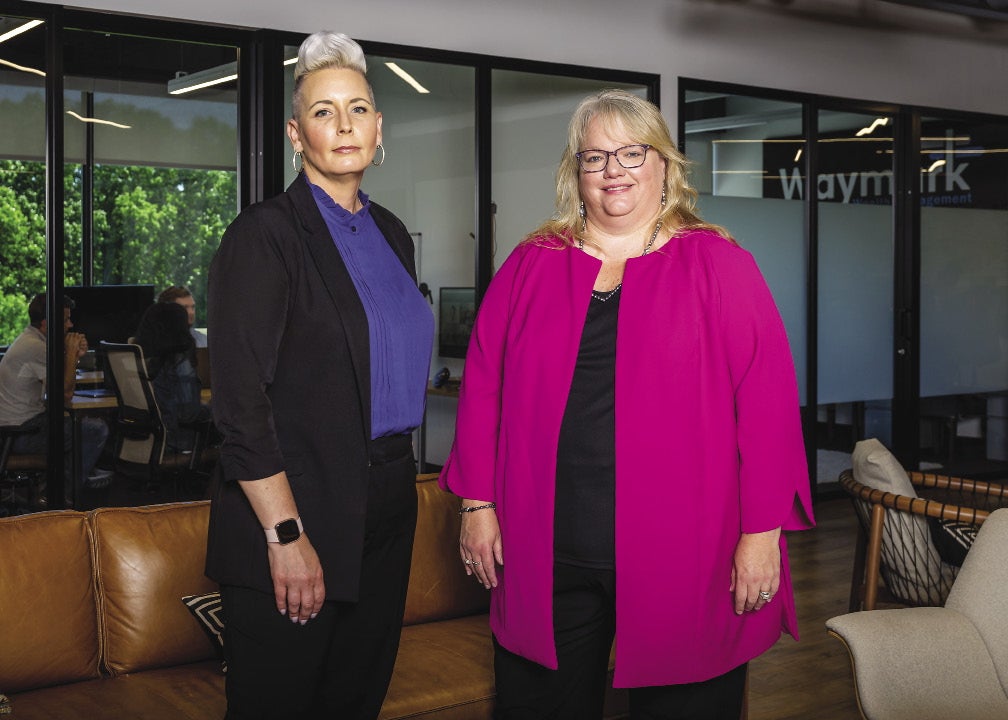 A woman with a white pompadour, blue blouse, and black jacket and pants stands next to a woman with blonde hair, glasses, pink jacket, and a black shirt and pants.
