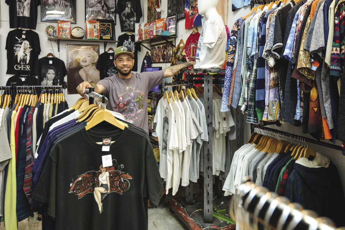 A man in a baseball cap and grey shirt stands among racks of t-shirts in front and beside him.