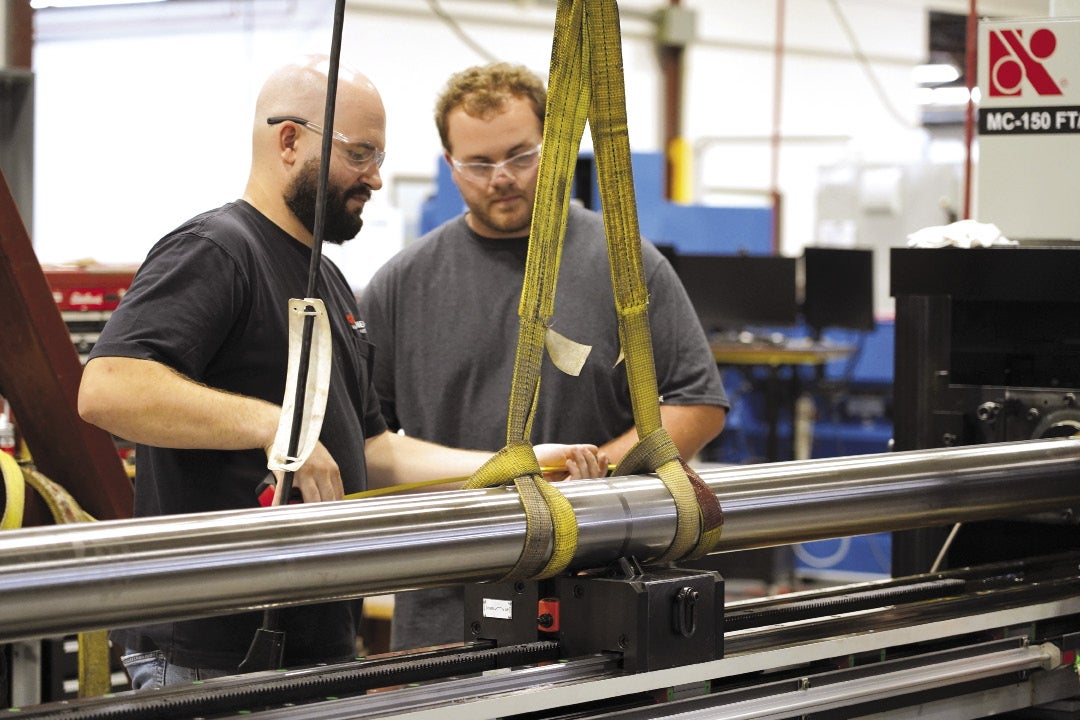 Two workers lower a rolled piece of steel onto a machine.